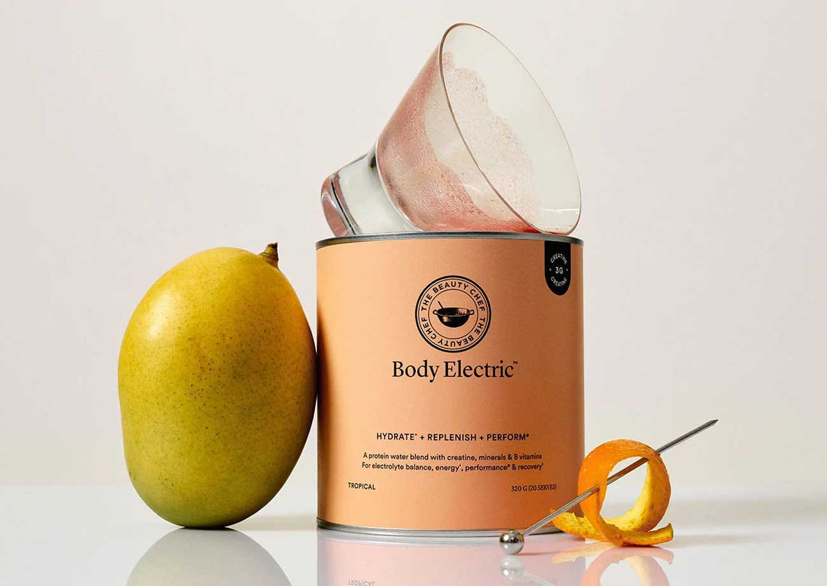 A tub of The Beauty Chef Body Electric Tropical with a fresh mango leaning against it, orange peel and a cocktail glass