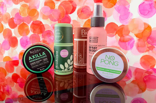 Five bicarb-free natural deodorants, including Axilla deodorant paste and No Pong, lined up on a glossy surface against a pink and orange watercolour dot background