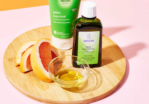 A dark green Weleda Birch oil bottle and green body scrub tube stand on a round wooden tray. Grapefruit wedges and a small glass bowl of golden oil sit in front on the pink backdrop.