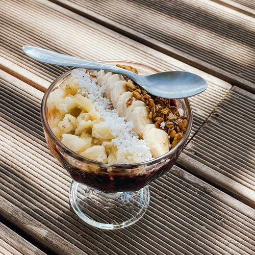 Chia pudding fills a clear stemmed glass, with banana slices, shredded coconut and granola piled on top. A metal spoon rests across the rim, and the glass stands on a slatted wooden table.