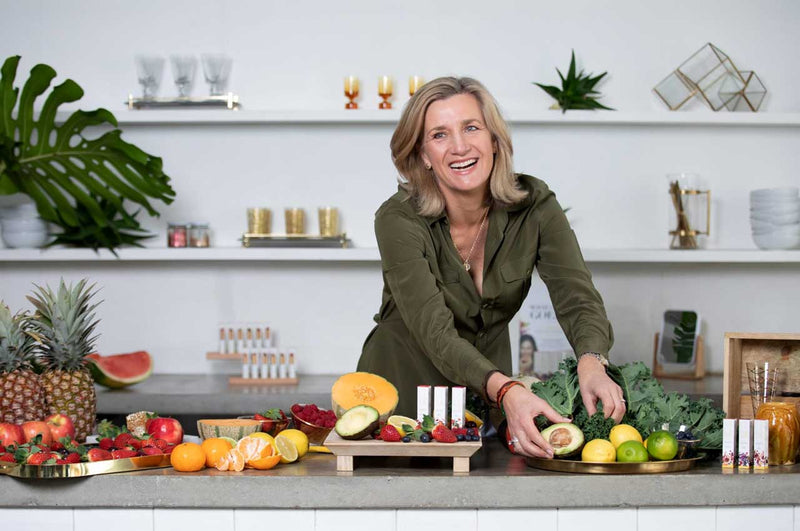 Food scientist Cindy Lüken smiling over a kitchen bench of fresh avocado, rockmelon, kale, citrus, strawberries and pineapples, with white lük beautifood lipstick tubes on a wooden stand in the middle
