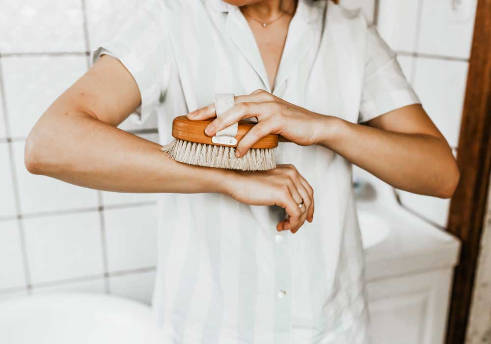 Women in a bathroom demonstrating how to use a dry body brush on her arm