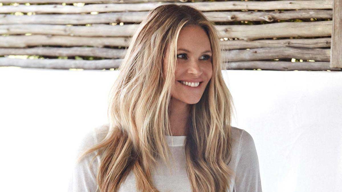 Elle Macpherson smiling, with long wavy blonde hair and a white top, in front of a rustic timber wall