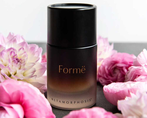 A cylindrical Formë Metamorphosis perfume bottle stands among loose pink flowers and petals. Its dark upper section fades to amber near the base, with the product name printed across the front.