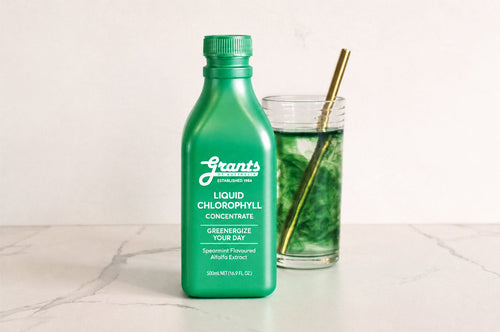 A green bottle of Grants Liquid Chlorophyll Concentrate next to a glass of green chlorophyll water with a gold straw on a white marble bench