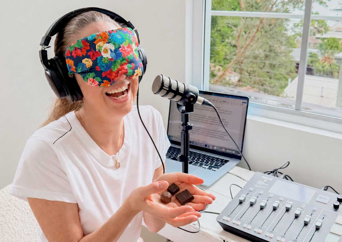 Irene Falcone laughing in a colourful floral blindfold and headphones, holding pieces of chocolate during a blind taste test at a podcast microphone