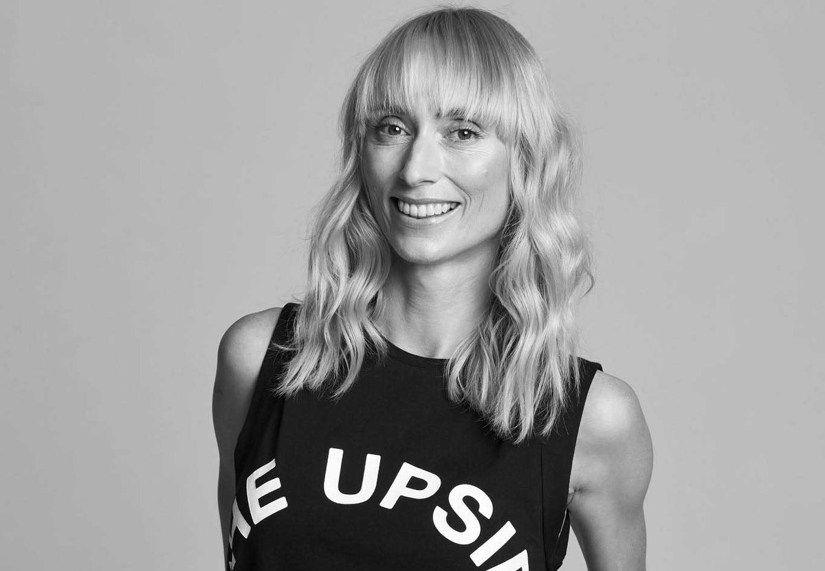 Black and white portrait of yoga teacher Kate Kendall smiling, with long wavy blonde hair and a fringe, wearing a sleeveless black tank top