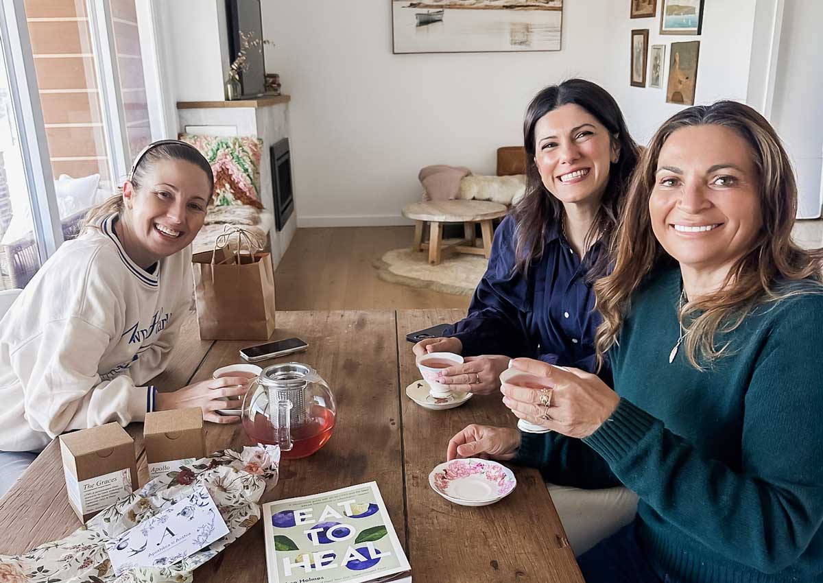 Irene Falcone, Lee Holmes and naturopath Anthia Koullouros smiling over cups of tea at a timber table with a glass teapot and a copy of Lee's book Eat to Heal