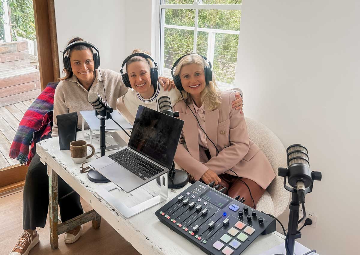 Lee Holmes, Irene Falcone and Amy Down in a pink blazer smiling in headphones around the podcast desk, with a laptop, microphones and a mixing desk in the bright cabin studio