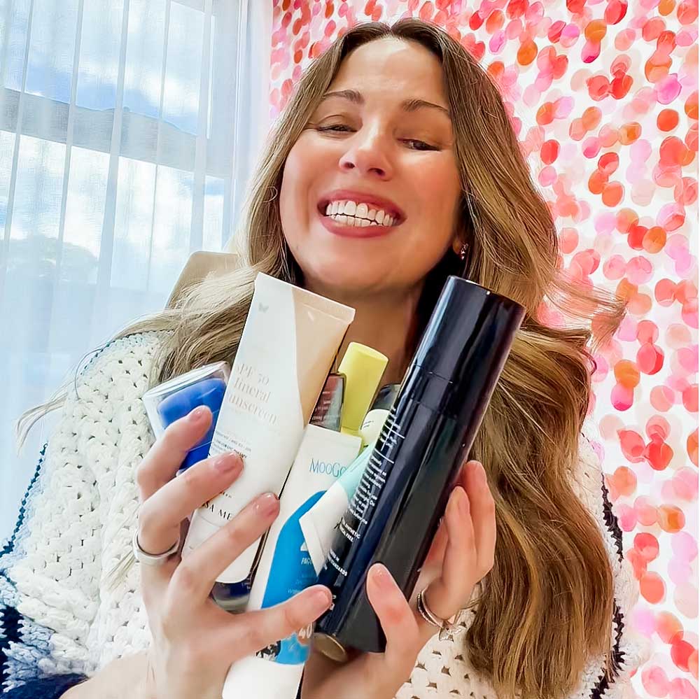 Irene Falcone smiling and holding an armful of mineral face sunscreens in front of a pink and orange watercolour dot wall