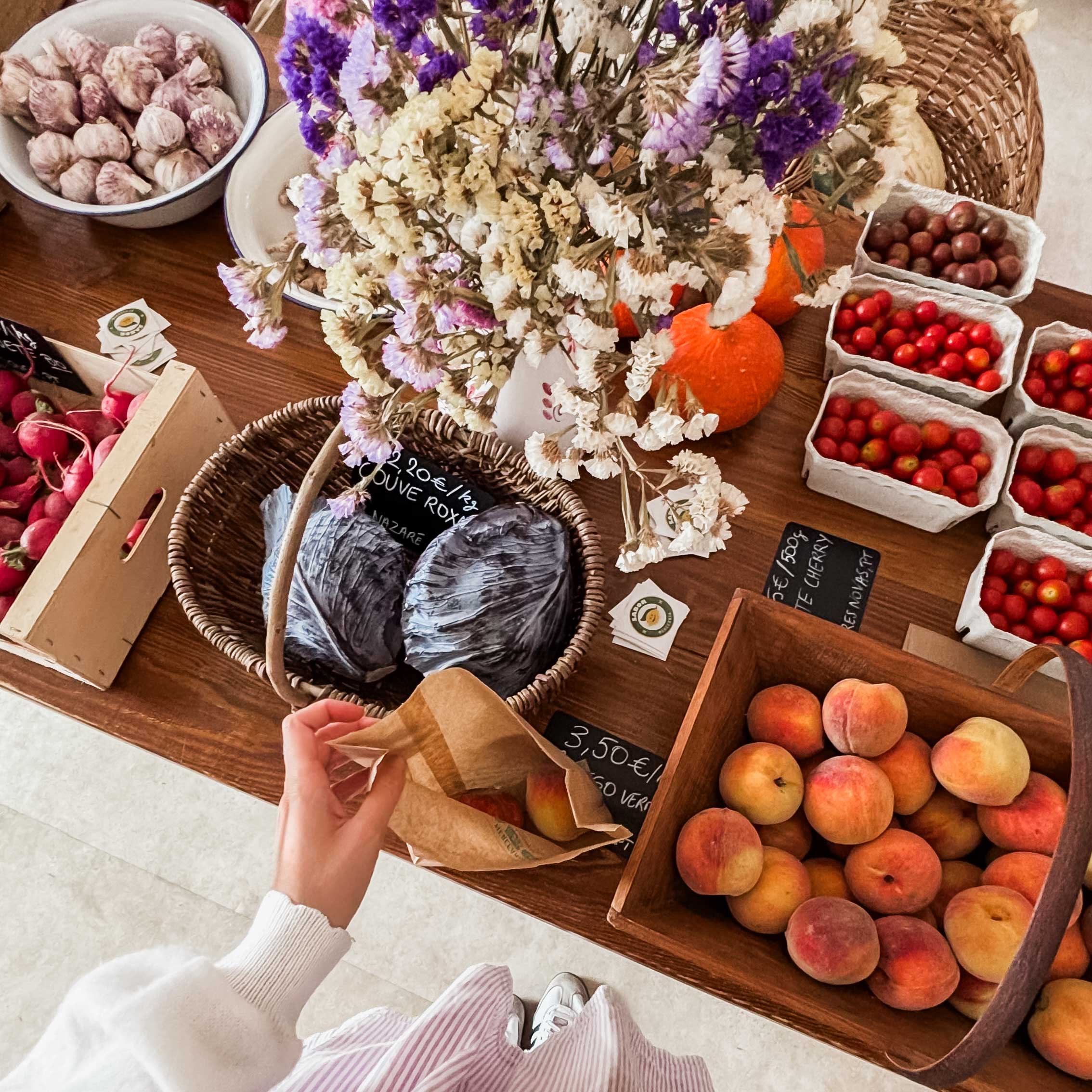A person reaches into a paper bag while shopping at a rustic farmers market stall filled with fresh peaches, tomatoes, cherries, red cabbage, garlic, pumpkins and dried flowers displayed in wooden crates and baskets.