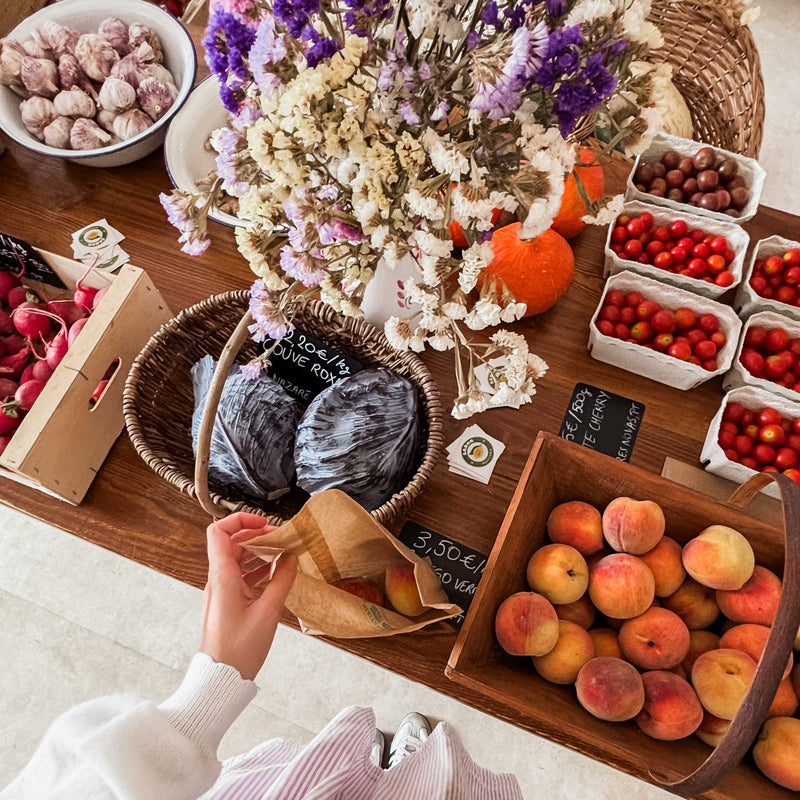 A person reaches into a paper bag while shopping at a rustic farmers market stall filled with fresh peaches, tomatoes, cherries, red cabbage, garlic, pumpkins and dried flowers displayed in wooden crates and baskets.