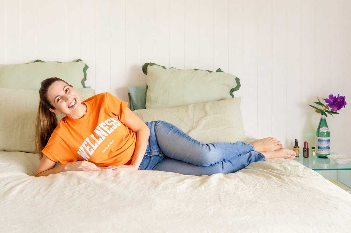 Irene Falcone smiling and lying on her side on a bed with cream linen and green-edged pillows, wearing an orange Wellness t-shirt and jeans, with a purple orchid and small bottles on the glass bedside table