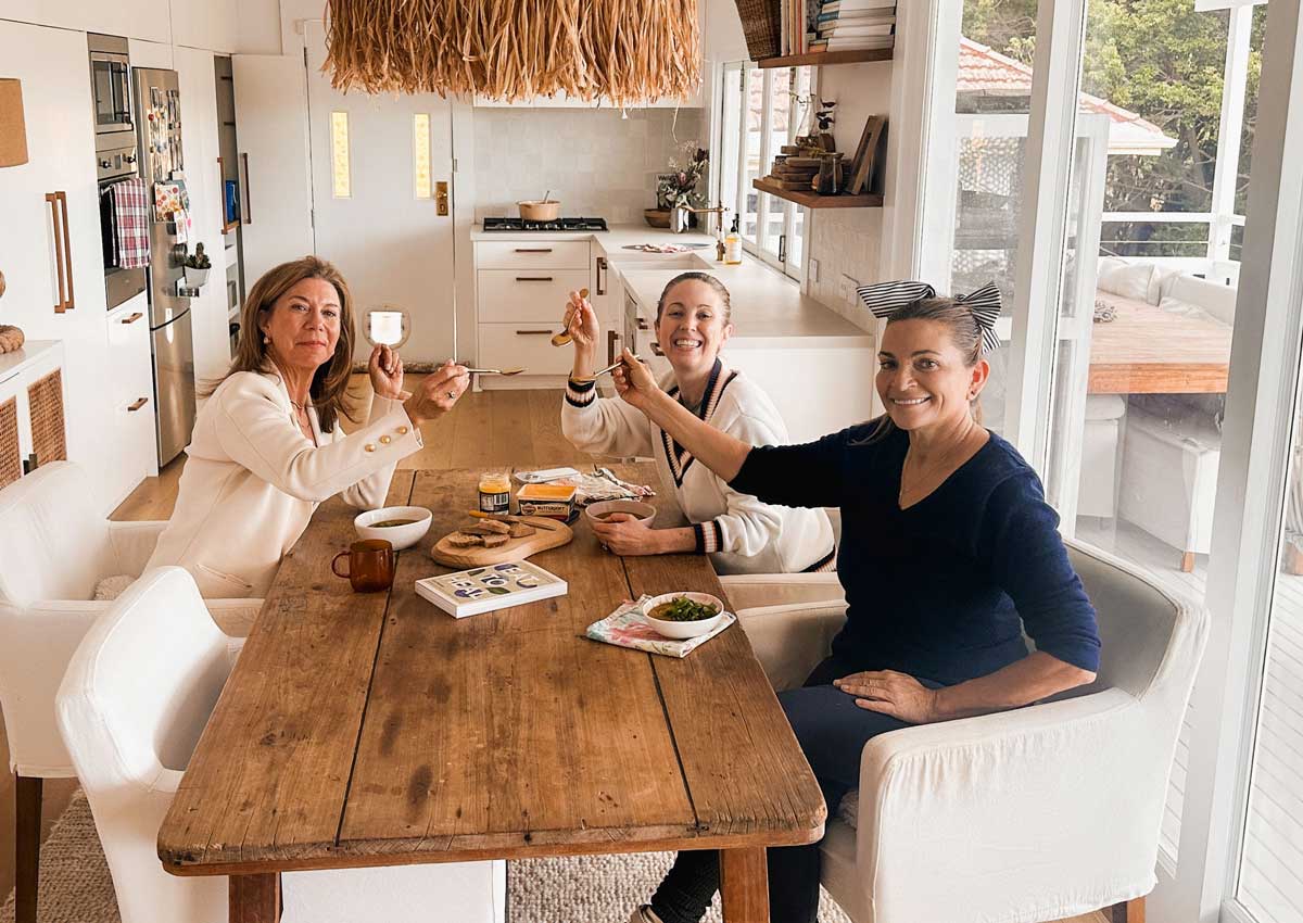 Michele Chevalley-Hedge, Lee Holmes and Irene Falcone smiling and raising small cups at a long timber dining table with snacks and books, in a bright white kitchen with a woven pendant light