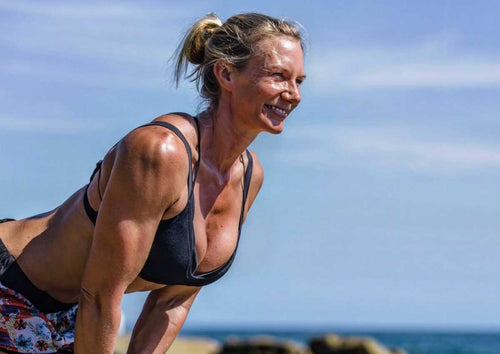 Strength coach Alexa Towersey laughing in a black sports bra and floral leggings, leaning forward mid workout on the rocks by the ocean under a blue sky