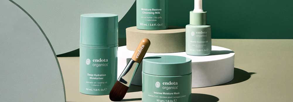 A collection of Endota Organics skincare products in green packaging, including moisturizers, a mask, a serum, and a brush, arranged on cylindrical display blocks against a green background.