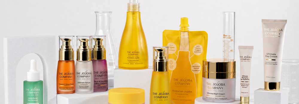 A collection of Jojoba skincare products, including creams, serums, and cleansers in white and gold packaging, arranged on and around textured beige blocks.