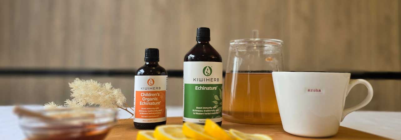 Two bottles of Kiwiherb Echinature stand behind a glass teapot and a white cup labeled aroba, with sliced lemons and a blurred jar of honey in the foreground.