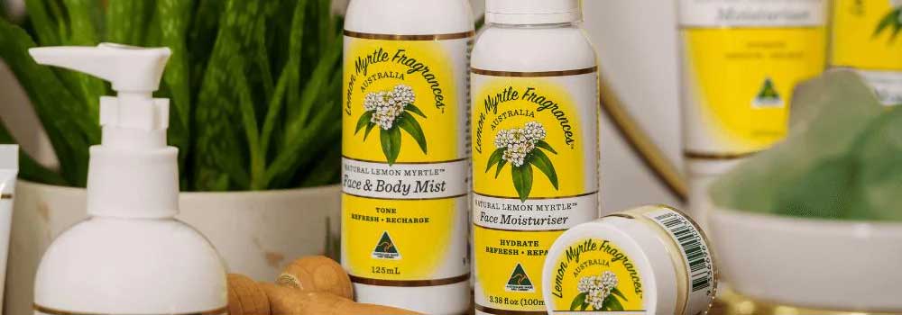 Several skincare products with yellow labels and white lids are displayed on a counter, featuring “Lemon Myrtle Fragrances” branding. A green plant is visible in the background.