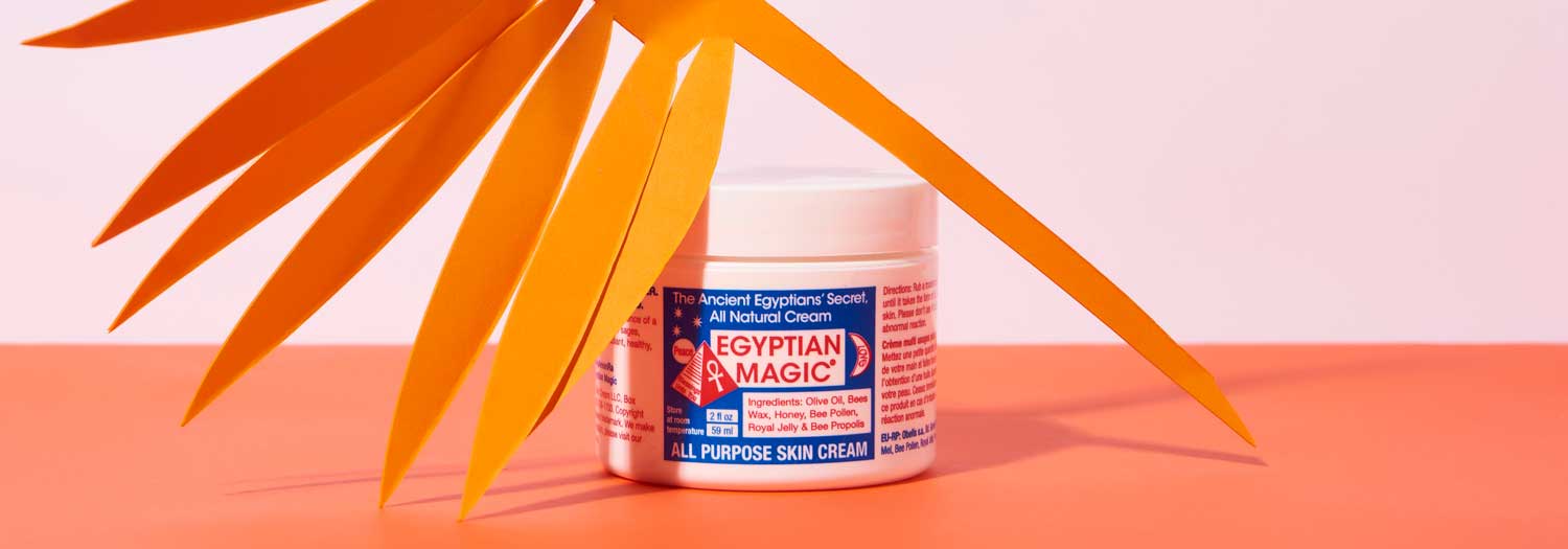A jar of Egyptian Magic All Purpose Skin Cream sits on an orange surface with a cutout of orange palm leaves casting a shadow over it.