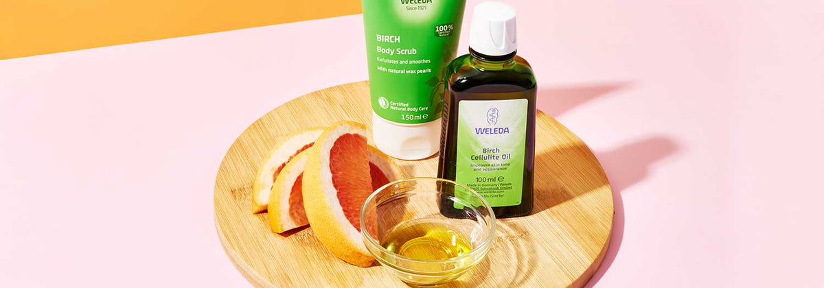 A wooden tray with grapefruit slices, a small bowl of oil, a tube of Weleda Birch Body Scrub, and a bottle of Weleda Birch Cellulite Oil, on a pink and yellow background.