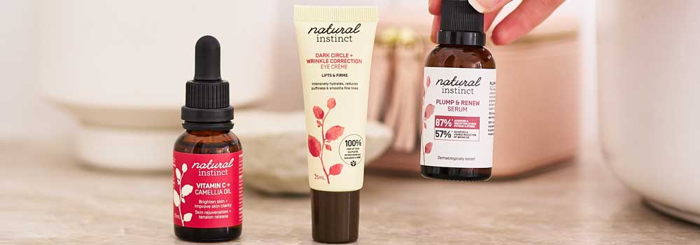 Three Natural Instinct skincare products, including Vitamin C & Camellia Oil, Eye Cream, and Plump & Protect Serum, are displayed on a beige surface with a hand picking up one bottle.