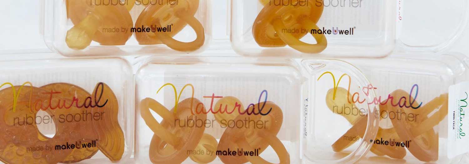 Several brown natural rubber baby pacifiers are displayed in clear plastic cases labeled Natural rubber soother and made by makeuwell. The cases are arranged in two rows.