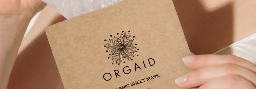 A person holds an Orgaid organic sheet mask packet, partially open to reveal the product inside. The packet features a black floral design on a brown background.