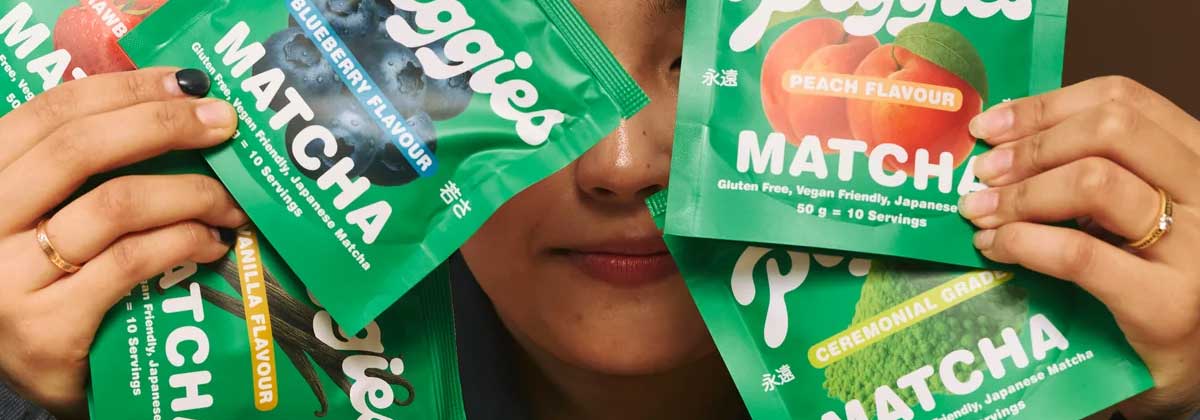 A person holds up four colorful packets of matcha powder in front of their face, with flavors including blueberry, peach, vanilla, and ceremonial grade.