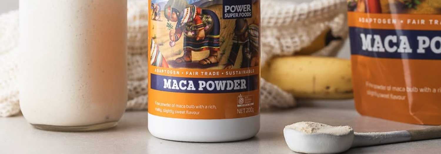 A container and a packet of maca powder are on a table with a spoonful of powder, a banana, and a glass of a creamy drink in the background. The product is labeled fair trade and sustainable.