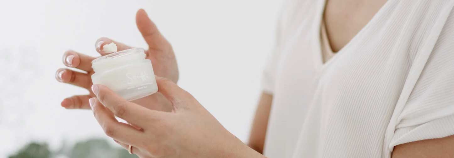 A person wearing a white shirt holds a small jar of cream in one hand and uses the other hand to scoop some cream out with their finger.