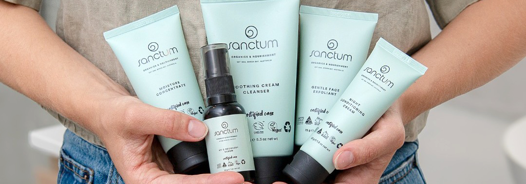 Person holding five Sanctum skincare products, including tubes and a bottle, with light green packaging and black caps, against a neutral background.