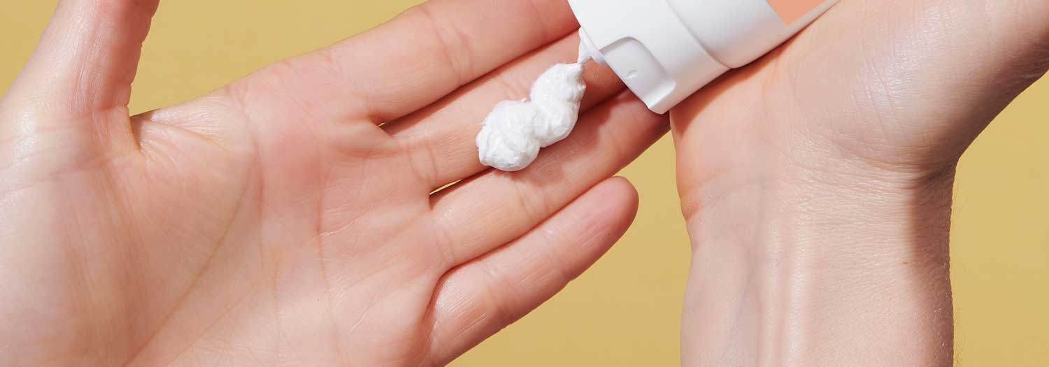 A person squeezes a white cream or lotion from a tube onto the palm of their hand, with a yellow background.