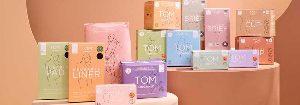 Various TOM Organic feminine hygiene products, including reusable pads, liners, briefs, tampons, and menstrual cups, are displayed in colorful boxes on beige curved platforms.