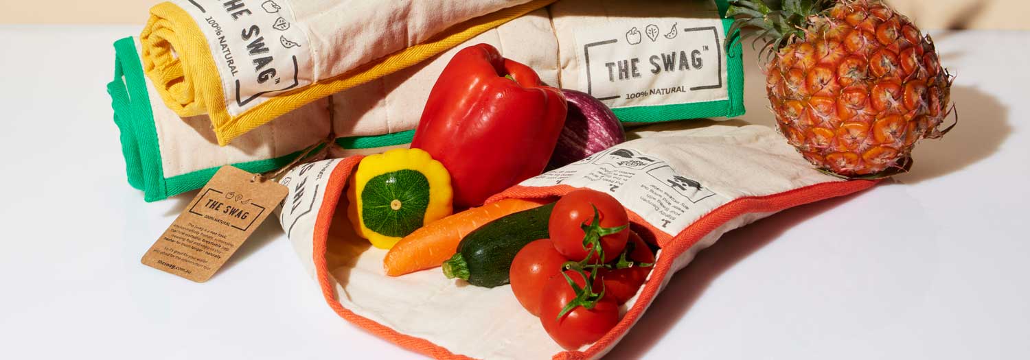 Reusable produce bags filled with fresh vegetables—tomatoes, carrot, zucchini, capsicum, and a red onion—sit on a white surface next to a whole pineapple and other rolled-up bags.