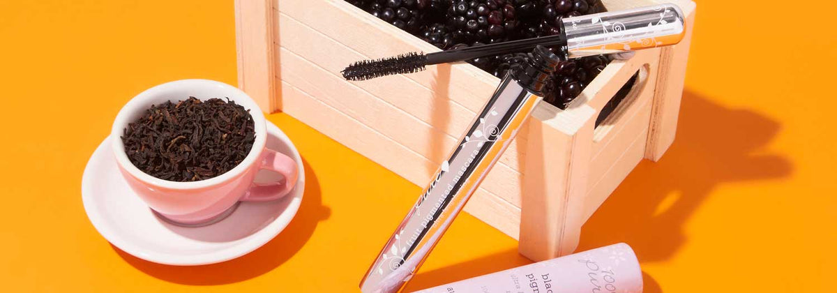 A pink cup filled with loose tea sits next to a wooden crate of blackberries and an open mascara tube, all arranged on a bright orange surface.