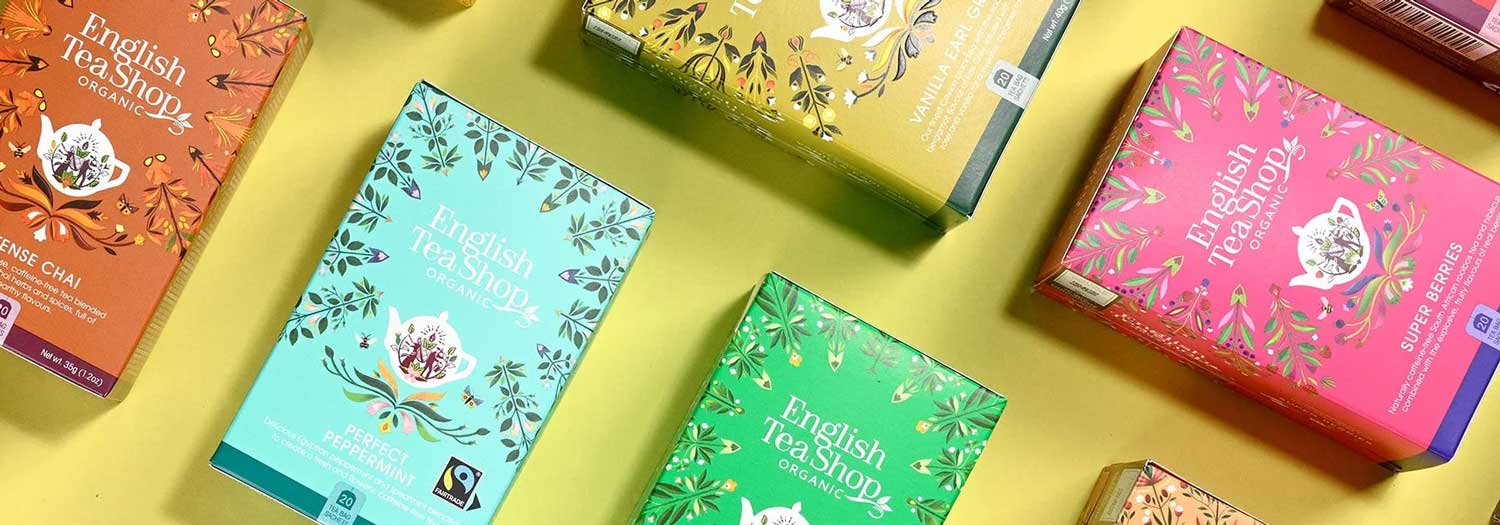 Assorted colorful boxes of English Tea Shop organic teas are arranged diagonally on a yellow background, displaying various flavors and vibrant packaging designs.