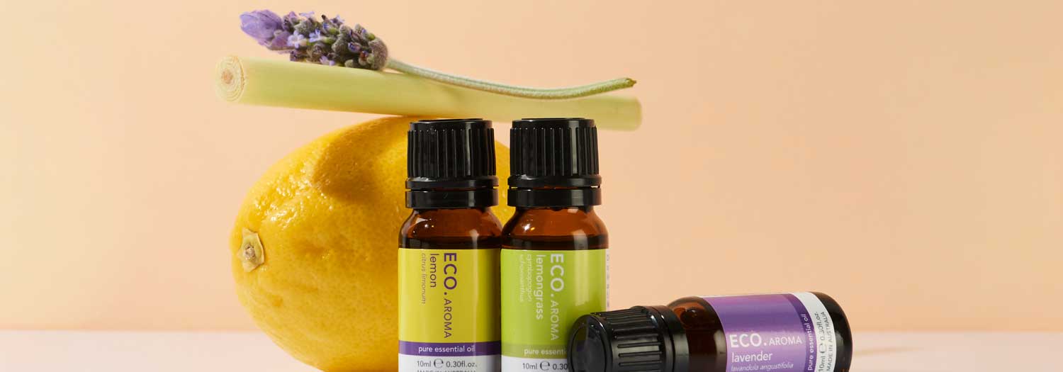 Three essential oil bottles labeled Eco Aroma are displayed with a lemon, a sprig of lavender, and a stalk of lemongrass on a light peach background.