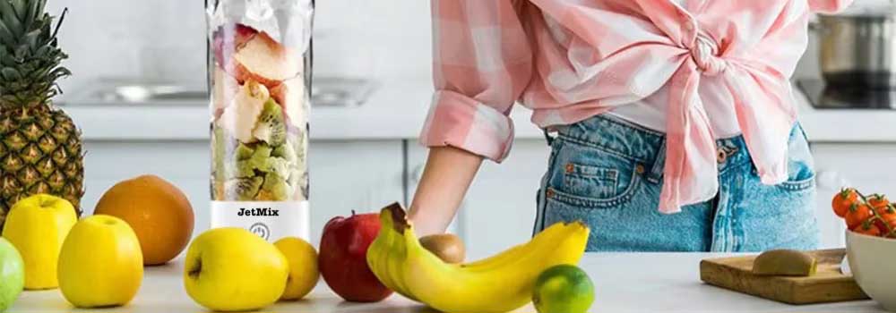 A person in a pink plaid shirt stands in a kitchen next to a JetMix blender filled with fruit, surrounded by apples, bananas, a pineapple, and other fresh produce on the counter.