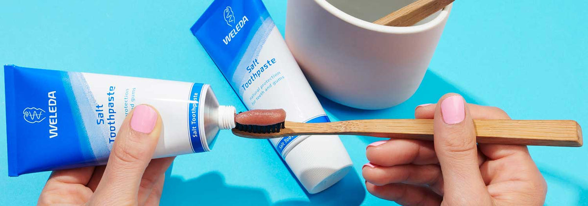 A person squeezes brown Weleda salt toothpaste onto a wooden toothbrush over a blue surface, with another toothpaste tube and a white cup in the background.
