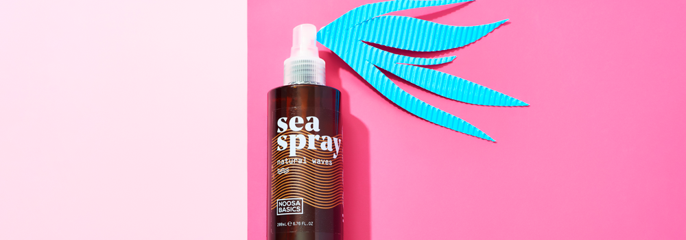 A bottle of sea spray hair mist with a blue paper cutout resembling a splash on a pink and light pink background. The label reads sea spray innovative wave and Noosa Basics.