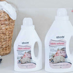 Two bottles of Abode Fabric Softener Comfort Fresh on a white laundry bench beside a woven basket and folded towels