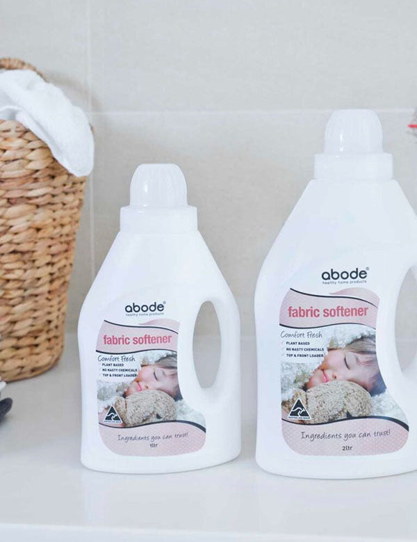 Two bottles of Abode Fabric Softener Comfort Fresh on a white laundry bench beside a woven basket and folded towels