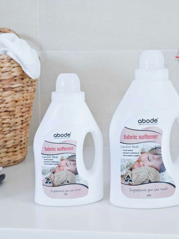 Two bottles of Abode Fabric Softener Comfort Fresh on a white laundry bench beside a woven basket and folded towels