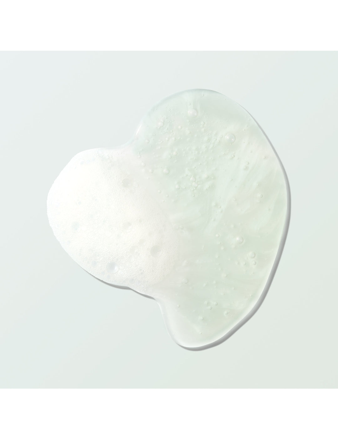 A clear Coconut Shower Gel swatch forms an irregular rounded pool on a pale mint surface. Tiny bubbles and a patch of white foam are visible within the gel.