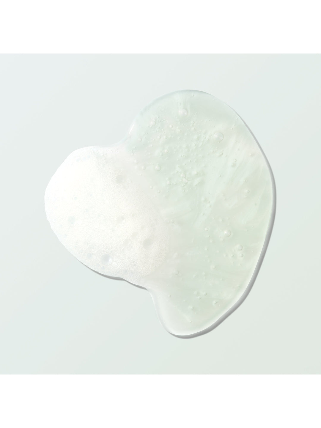 A clear Coconut Shower Gel swatch forms an irregular rounded pool on a pale mint surface. Tiny bubbles and a patch of white foam are visible within the gel.
