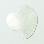 A clear Coconut Shower Gel swatch forms an irregular rounded pool on a pale mint surface. Tiny bubbles and a patch of white foam are visible within the gel.
