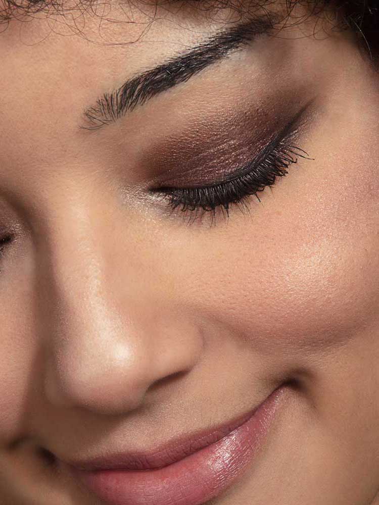 Close-up of a smiling person with closed eyes, smooth skin, shimmery brown eyeshadow from the 100% Pure Berry Naked Palette 9g, black eyeliner, long lashes, and naturally shaped brows.