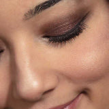 Close-up of a smiling person with closed eyes, smooth skin, shimmery brown eyeshadow from the 100% Pure Berry Naked Palette 9g, black eyeliner, long lashes, and naturally shaped brows.