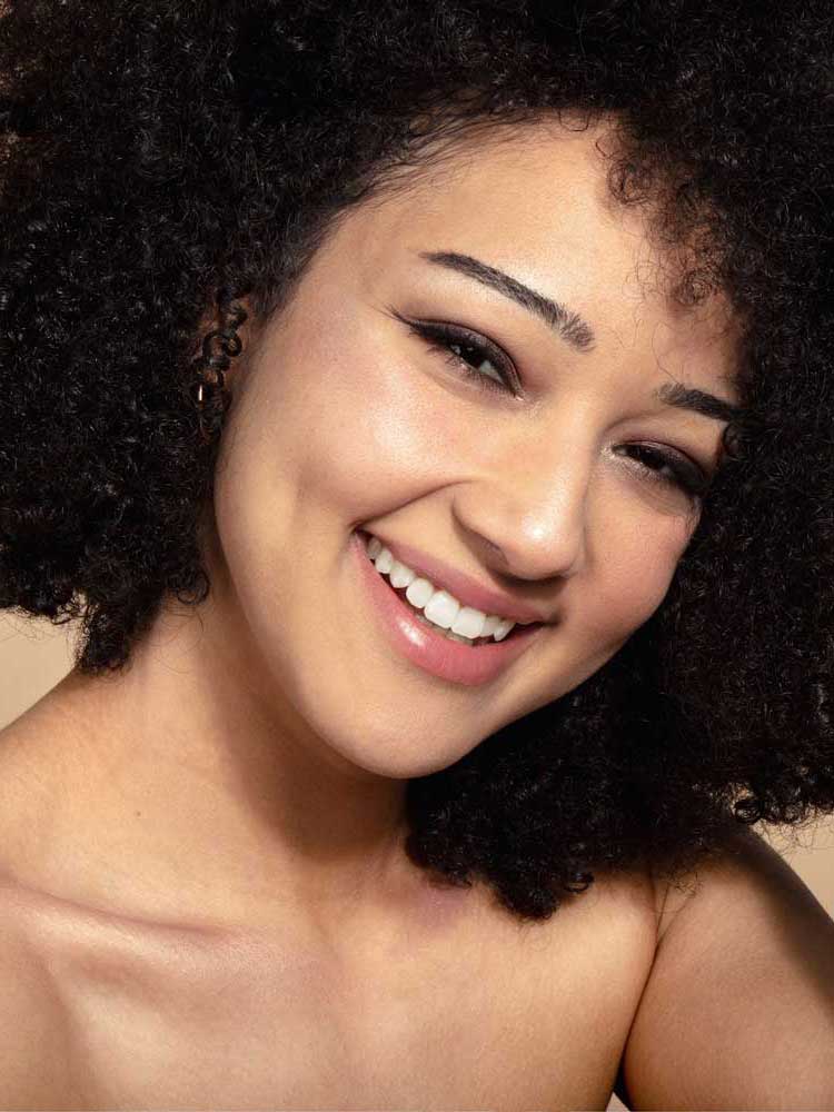 A young woman with curly dark hair smiles warmly at the camera, wearing natural makeup using shades from the 100% Pure Berry Naked Palette 9g, a vegan eyeshadow known for long-lasting wear, shown from the shoulders up against a neutral background.